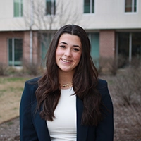 A young woman smiling outdoors, wearing a navy blazer over a white top, with a neutral background of buildings and bushes.