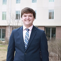 A young man in a suit and tie stands smiling outdoors in front of a modern building.