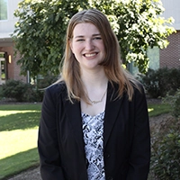 Young woman in a professional outfit smiling outdoors in front of a building and trees.