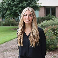 A young woman with long blonde hair smiles while standing outdoors in a professional setting.