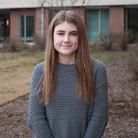 A young woman with long brown hair is standing outdoors, wearing a cozy gray sweater and smiling at the camera.