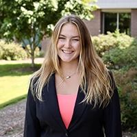 A smiling young woman in a business suit stands outdoors, with greenery in the background.