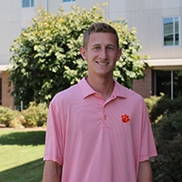 Young man in a pink polo shirt with a Clemson University logo, smiling outdoors in front of greenery.