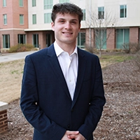 A young man in a dark blazer stands confidently outdoors, smiling in front of a modern building.