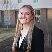 Professional headshot of a young woman in a black suit with long blonde hair, smiling outdoors.