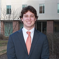 A smiling young man in a suit and orange tie standing outside a building.