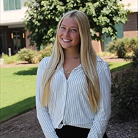 A smiling young woman with long blonde hair, wearing a white striped blouse and standing outdoors in a green environment.