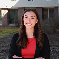 Young woman with long brown hair, wearing a red shirt and black cardigan, smiling confidently outdoors.