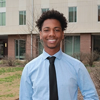 Smiling young man in a light blue dress shirt and black tie, standing outdoors in front of a modern building.