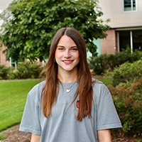 A young woman with long brown hair smiles while standing outdoors in front of greenery and a building.