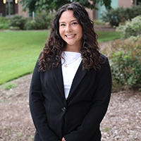 Professional headshot of a smiling young woman in a black blazer, standing outdoors in a landscaped area.