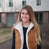A young woman with long hair smiles while wearing a brown vest in an outdoor setting.