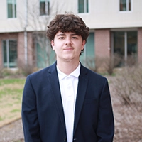A young man in business attire stands confidently outdoors, displaying a professional demeanor.