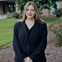 Professional portrait of a woman in a black blazer, smiling outdoors against a backdrop of greenery.