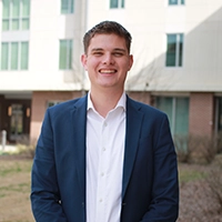 A professional headshot of a young man in a blazer standing outside a building, smiling confidently.