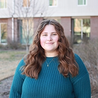Portrait of a woman with wavy, shoulder-length hair standing outside in a casual teal sweater.
