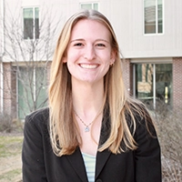 A smiling young woman in a black blazer stands outdoors with a modern building in the background.