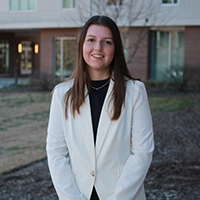 A young woman in a white blazer smiles confidently in an outdoor setting.