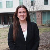 A professional headshot of a woman in a black blazer, smiling in front of a modern building.