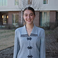Smiling young woman in a stylish gray blazer with decorative buttons standing outdoors in front of a modern building.