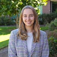 A young woman with long, wavy hair smiles while wearing a plaid blazer, standing in a garden setting.