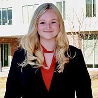 A professional portrait of a young woman in business attire, smiling and standing outdoors in front of a modern building.