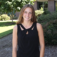 A young woman smiling while wearing a black sleeveless top, standing outdoors in a landscaped area.