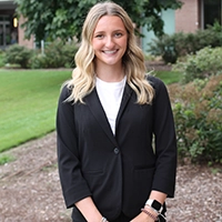 A professional young woman in a black blazer stands outdoors, smiling confidently in front of a green backdrop.