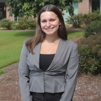 A smiling young woman in a professional grey blazer standing outdoors, with greenery in the background.
