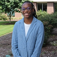 A smiling young woman wearing glasses and a blue blazer stands outdoors, surrounded by greenery.