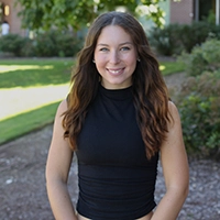 A smiling young woman with long brown hair wearing a black sleeveless top, standing outdoors in a grassy area.