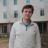 A young man wearing a light sweater stands in front of a building with large windows.