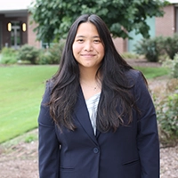 A young woman in a navy blazer stands outdoors with a green landscape and buildings in the background.