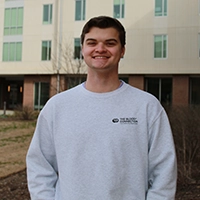 Smiling young man wearing a gray sweatshirt standing outside a building with large windows.