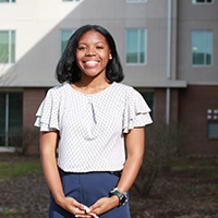 A smiling woman stands outdoors in front of a building, wearing a stylish blouse and navy pants.
