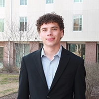A young man in a suit poses outdoors in front of a building, featuring curly hair and a confident expression.