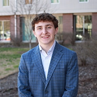 A young man in a blue checkered blazer standing outdoors, smiling confidently in front of a building.