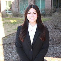 A young woman in a black blazer smiles confidently against a sunny outdoor backdrop.