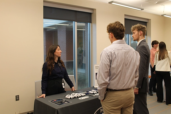 A professional woman speaks to two men at a recruitment event, with a table displaying promotional materials in the foreground.