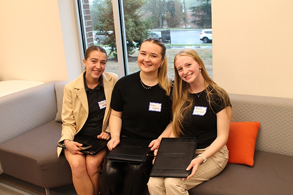 Three young women sit together on a couch, each holding a portfolio and smiling at the camera during a networking event.