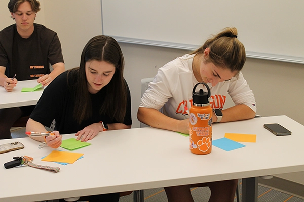Two students engaged in a collaborative activity, writing on colorful sticky notes at a classroom table. A Clemson-themed water bottle is visible in the foreground.