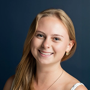 A smiling young woman with long blonde hair and gold earrings, set against a dark blue background.