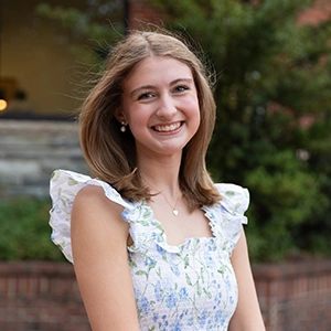 Smiling young woman in a white floral dress outdoors, with greenery and a brick walkway in the background.