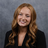 Studio headshot of a smiling person with long wavy red hair, wearing a black blazer against a gray backdrop.