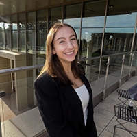 Smiling woman in a black blazer standing on an outdoor terrace beside a modern glass-fronted building.
