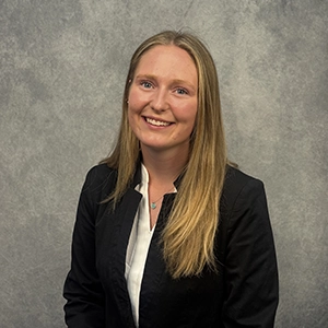 Studio portrait of a smiling woman with long blonde hair, wearing a black blazer over a white blouse against a gray backdrop.