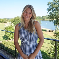 Smiling woman in a blue-and-white gingham dress standing by a railing with a lake and trees in the background.
