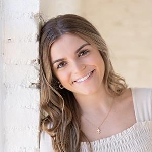 A smiling young woman with long wavy hair leans against a white brick wall, showcasing a warm and friendly demeanor.
