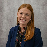 Smiling person with long red hair wearing a navy blazer and floral blouse against a gray studio backdrop.