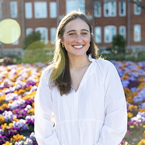 Smiling woman in a white blouse standing in front of colorful flower beds with a brick building in the background.